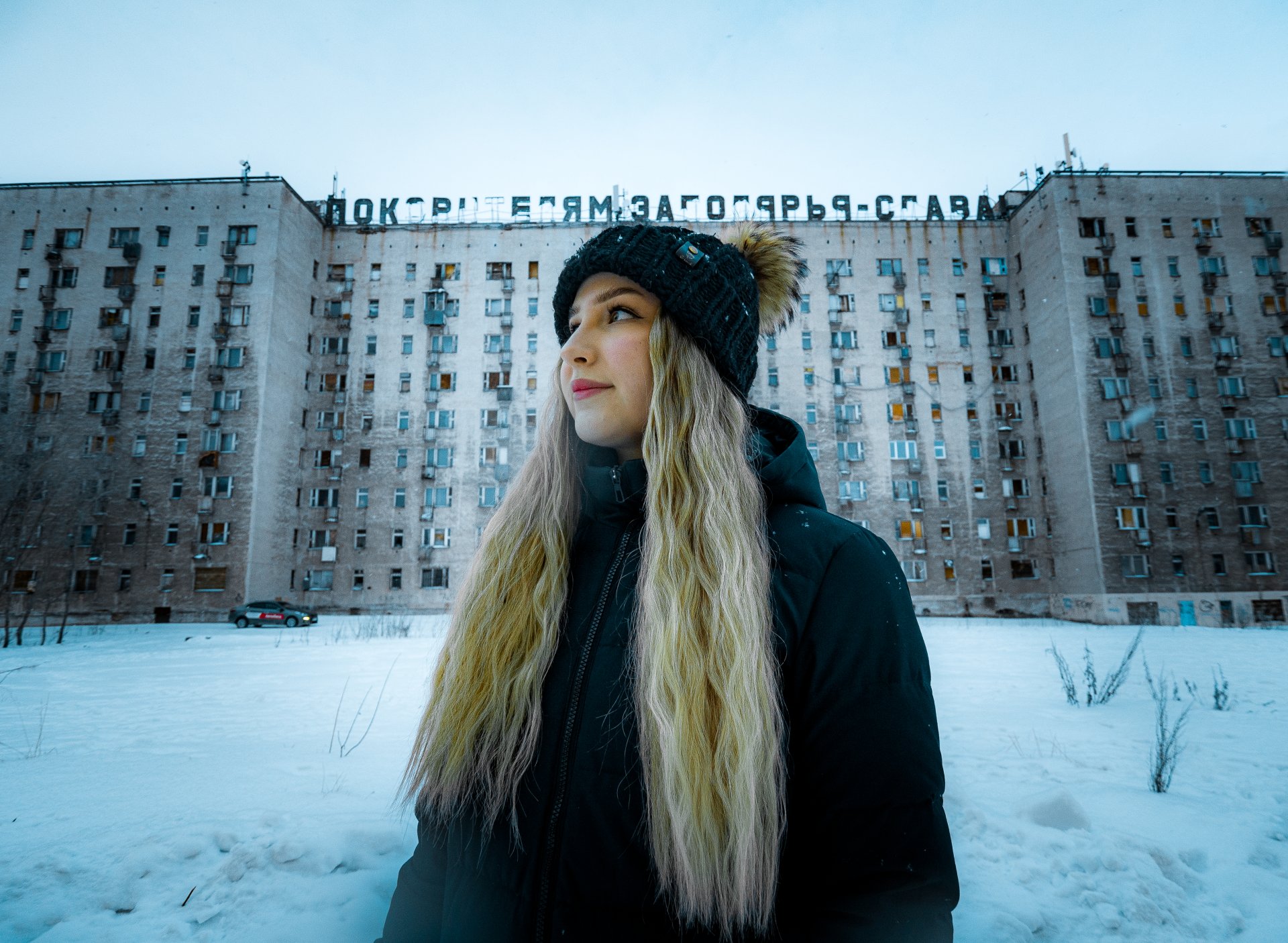 Karolina Anisimova - a local resident. In the background is one of the symbols of the city, a semi-abandoned house with a Soviet inscription on the roof: "Glory to the conquerors of the Arctic!"